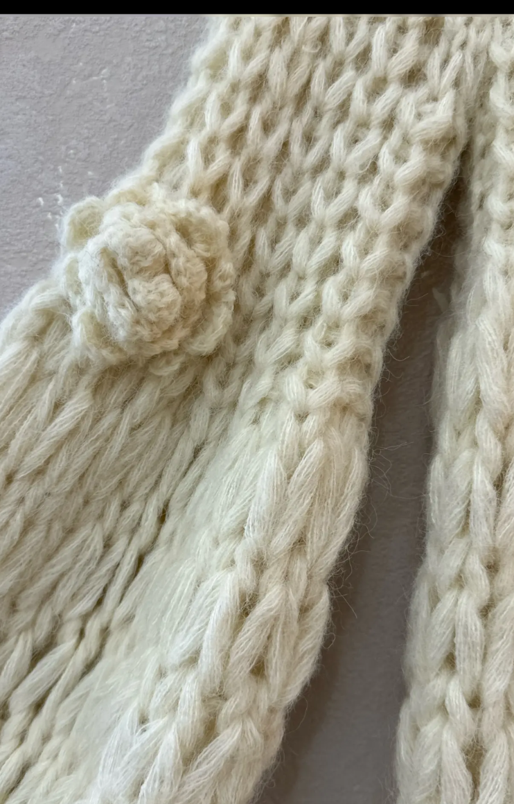 A close-up of a chunky knit cream-colored garment, showing a textured cable-knit pattern and a matching knitted pom-pom detail on the sleeve.