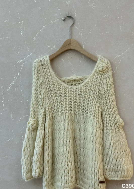 A cream-colored chunky knit sweater with balloon sleeves, hanging on a wooden hanger against a textured gray wall. Product details are printed on the bottom right of the image.