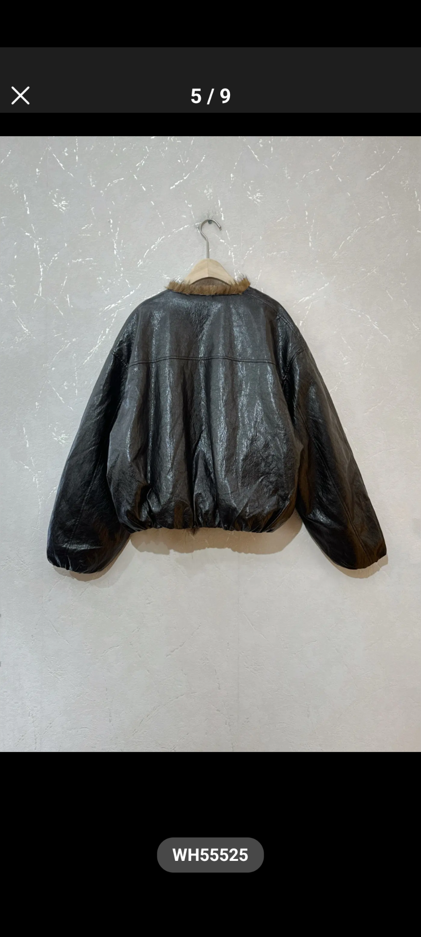 A black faux-leather bomber jacket with a tan faux-fur collar, hanging on a wooden hanger against a textured off-white wall.