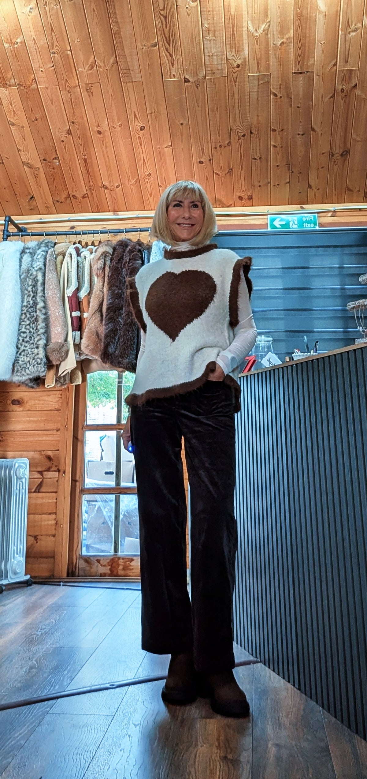 Maura beige heart sleeveless sweater layered over a white shirt with dark trousers and brown boots