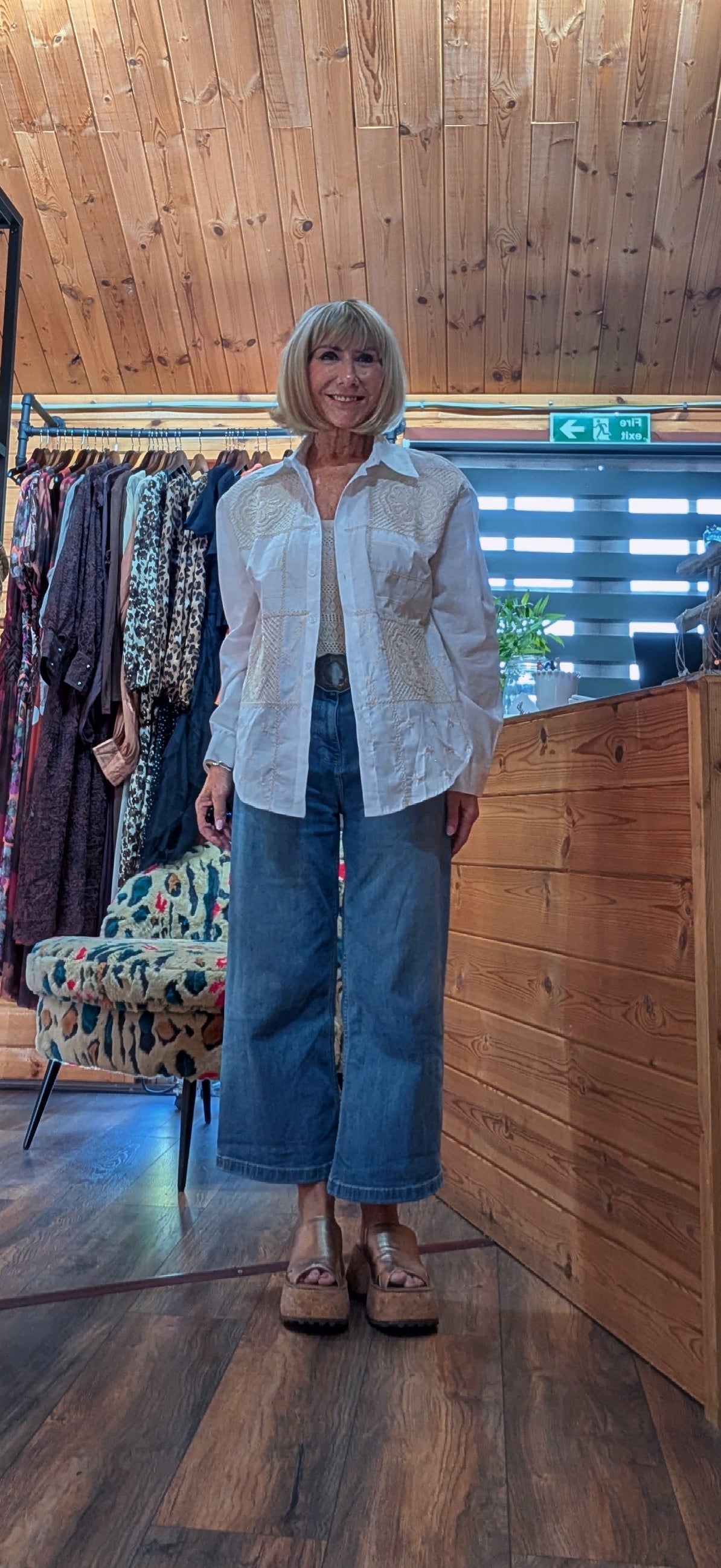 A woman with short blonde hair stands in a rustic wood-paneled boutique wearing a white long-sleeve button-up shirt and wide-leg blue jeans.