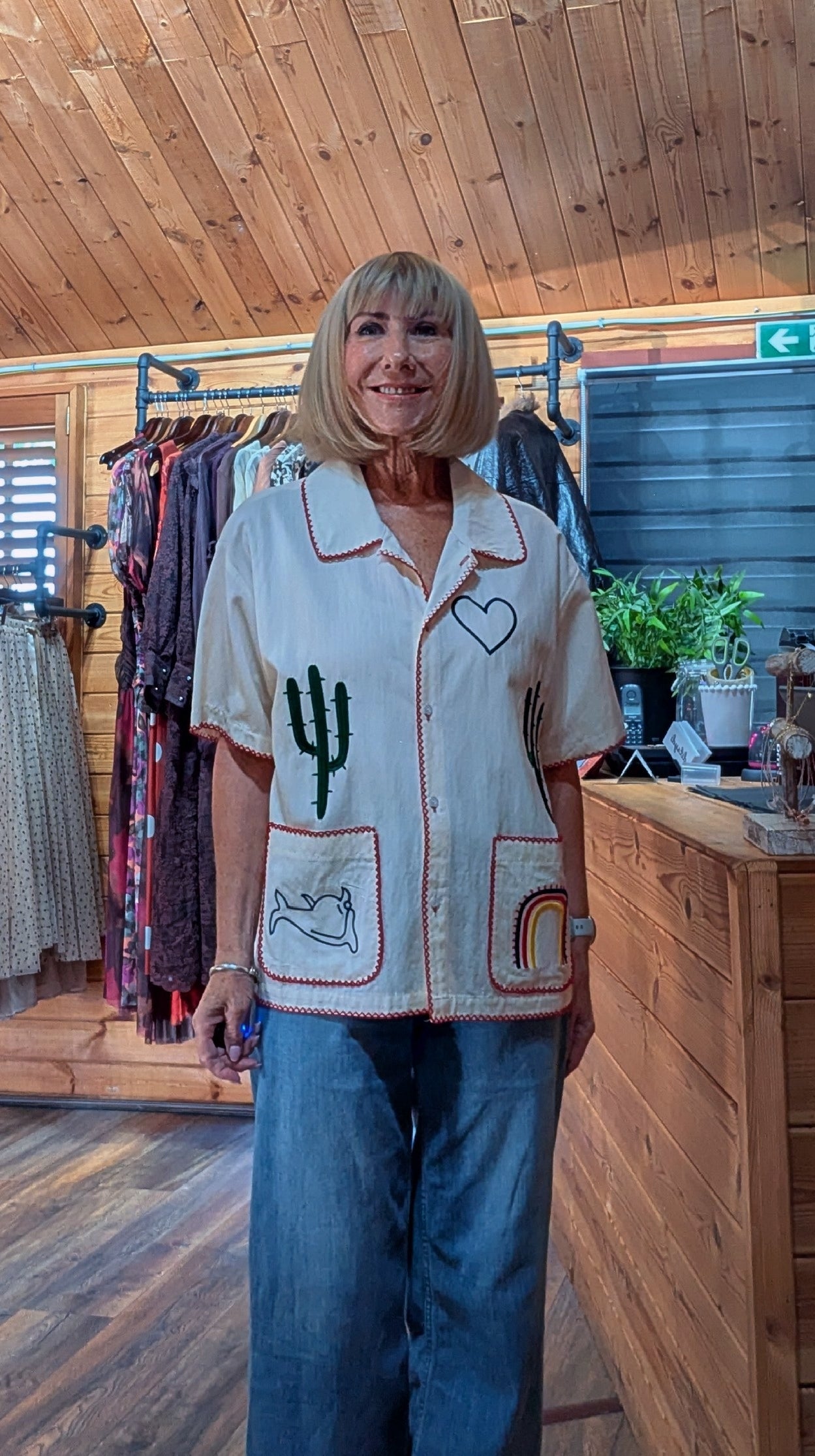 Ditsydi in the  shop wearing a cream-colored short-sleeve button-up shirt with embroidered desert motifs (cactus, heart, cat, rainbow) and blue jeans.