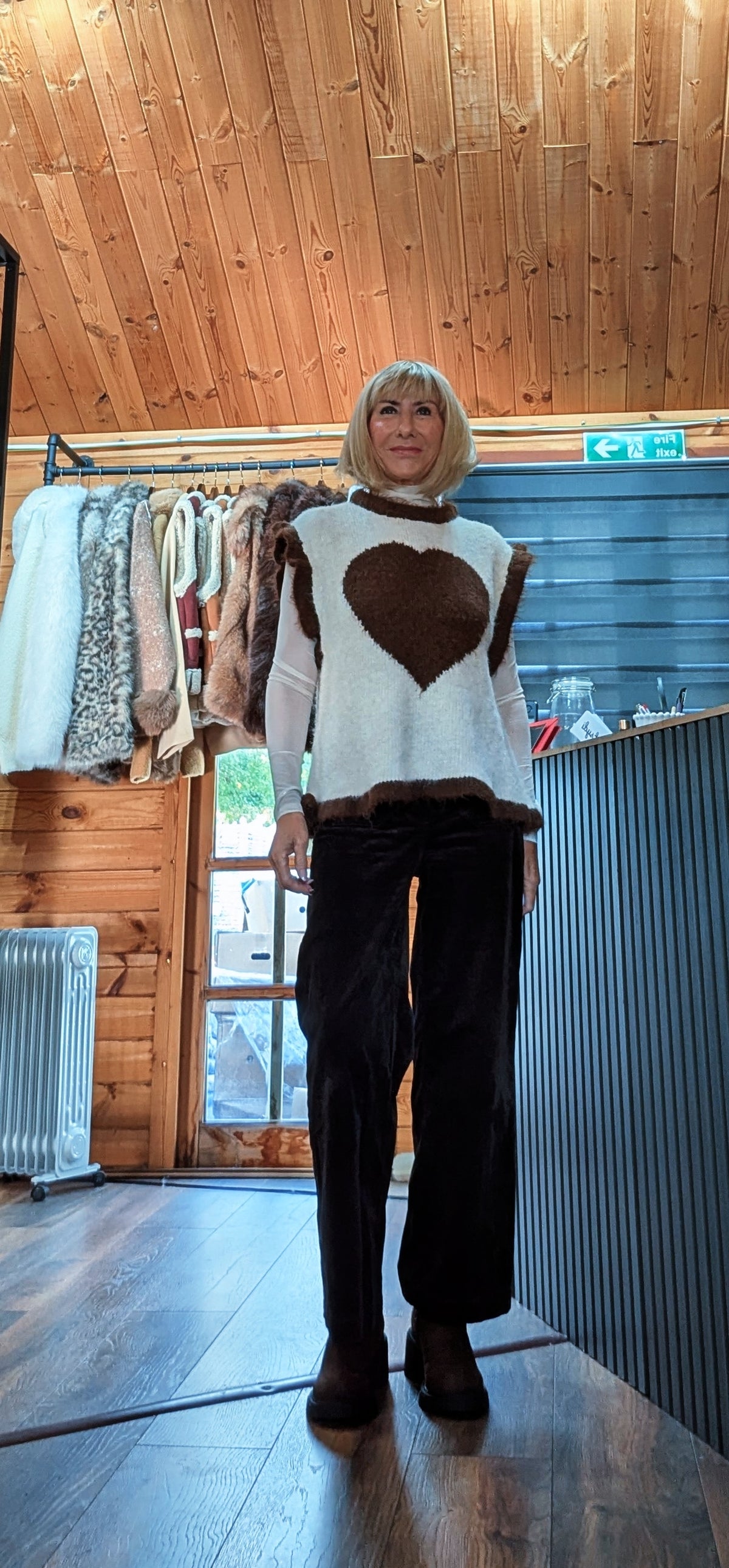 Maura beige heart sweater worn over a white shirt with dark wide-leg trousers