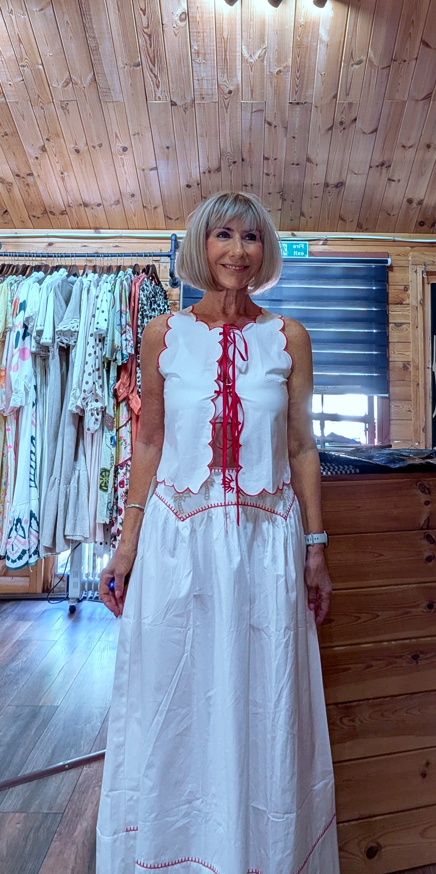 ditsydi UK modelling the Iris white cotton top and matching skirt inside a boutique, featuring red embroidery, scalloped edging and a flowing maxi skirt.