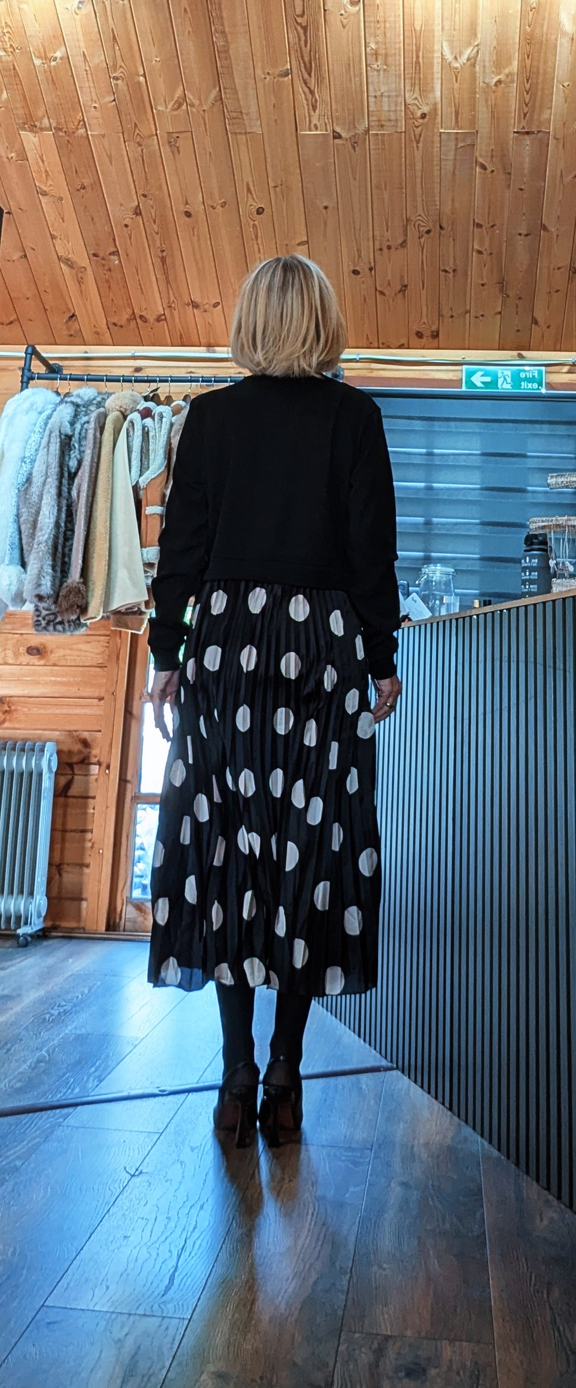 Back view of Hettie showing the plain black knitted top and pleated skirt with large white spots.