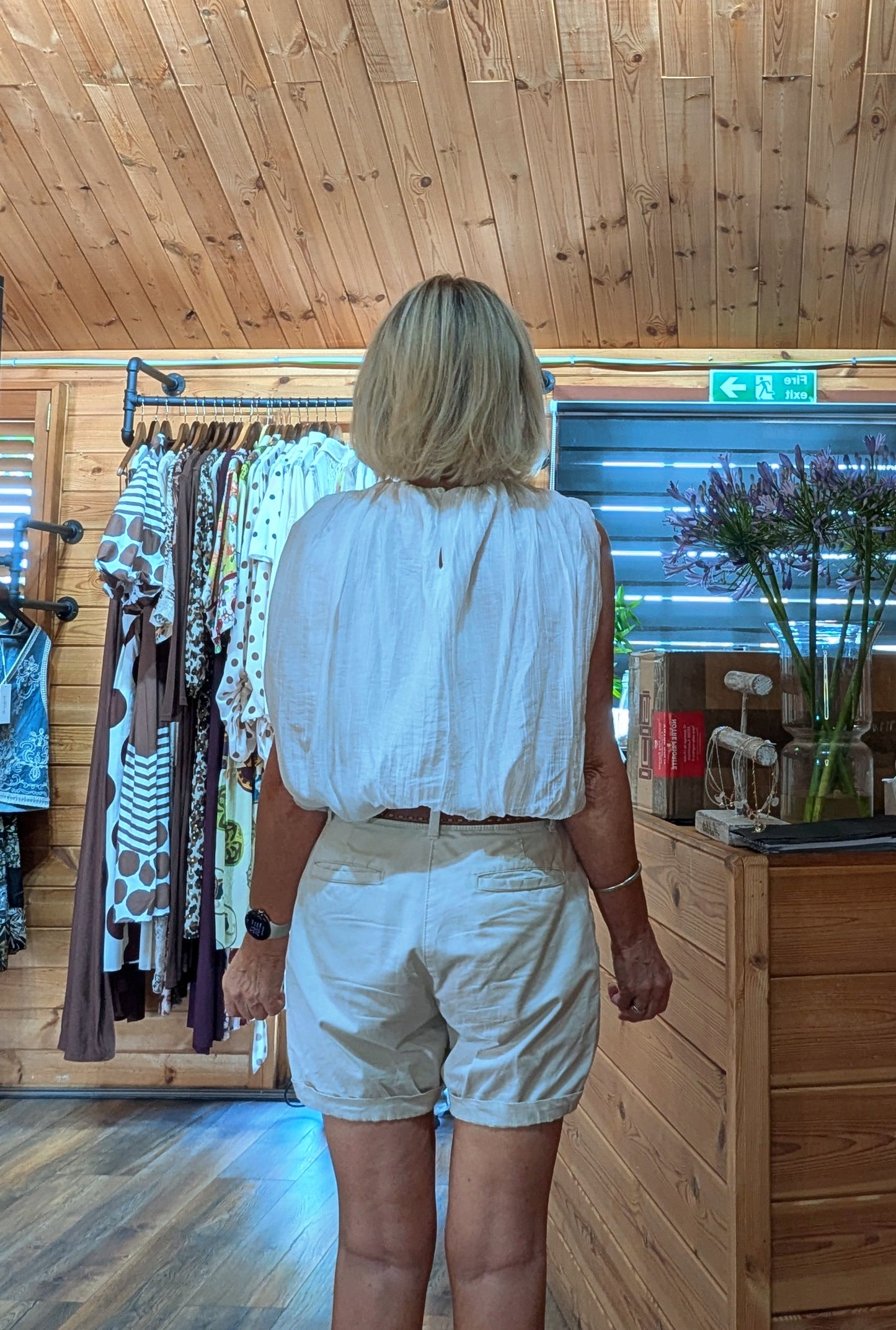 A woman viewed from behind wearing a white sleeveless cropped top and light beige shorts inside a wooden shop with clothing racks and a counter in the background.