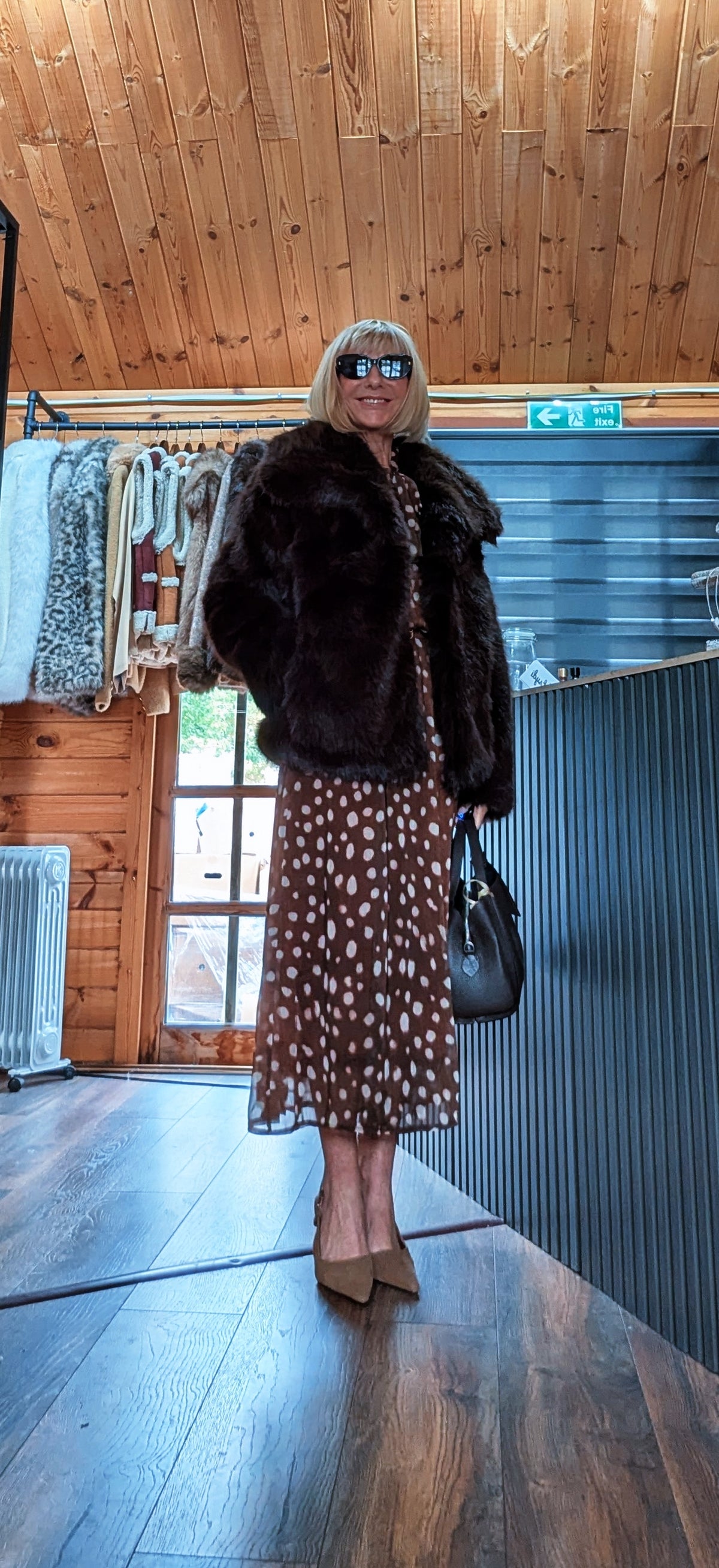 Angled view of Bella chocolate faux fur jacket worn over a spot-print dress with a black handbag