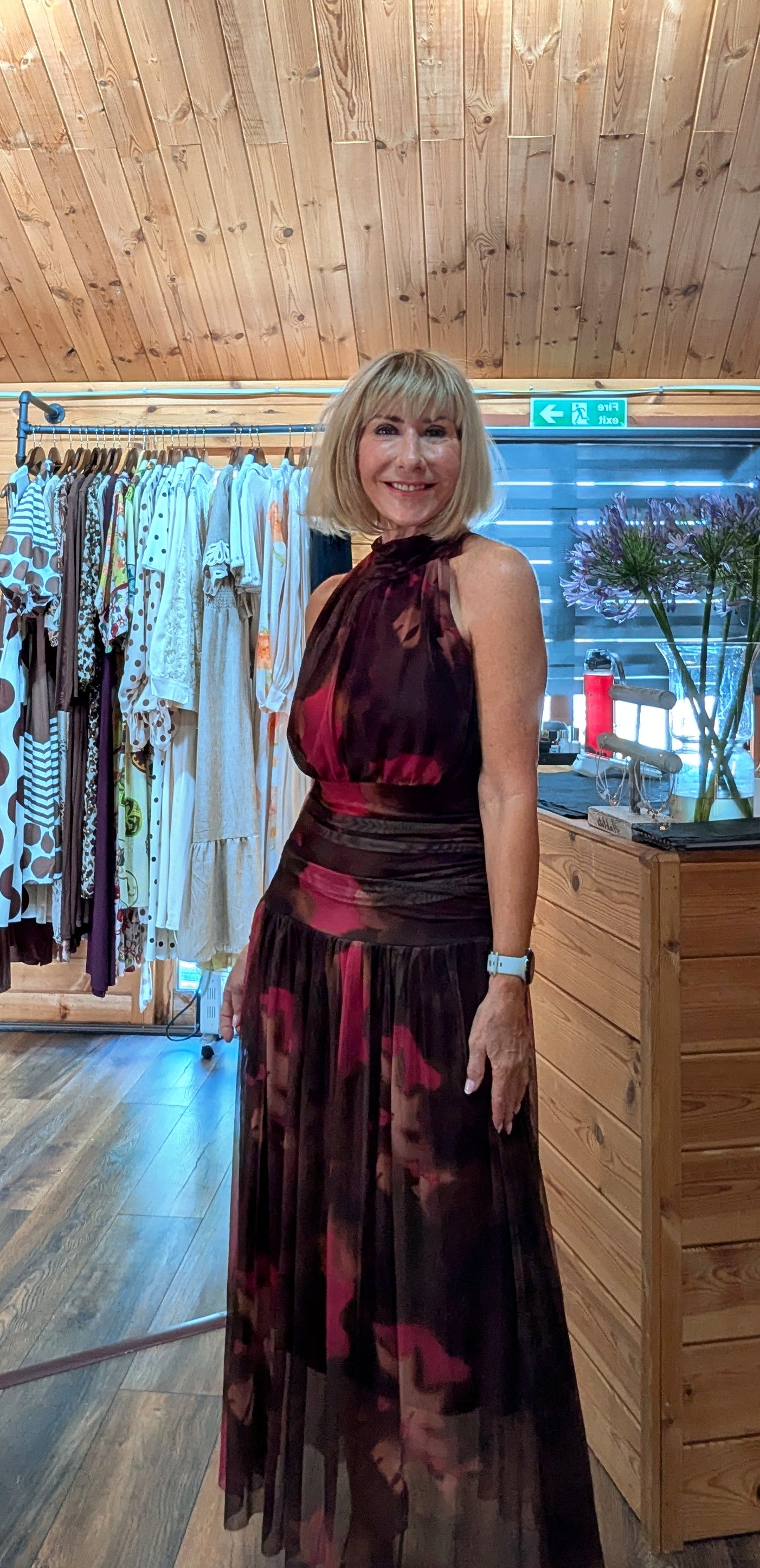 A woman with blonde hair wearing a dark red and burgundy patterned halter-neck maxi dress, standing inside a clothing boutique 