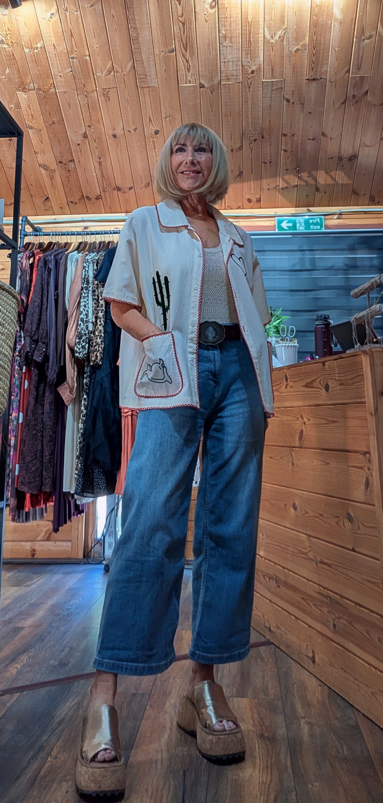 ditsydi in her boutique wearing a beige short-sleeve button-up shirt with a cactus patch, blue wide-leg cropped jeans, and brown platform sandals.
