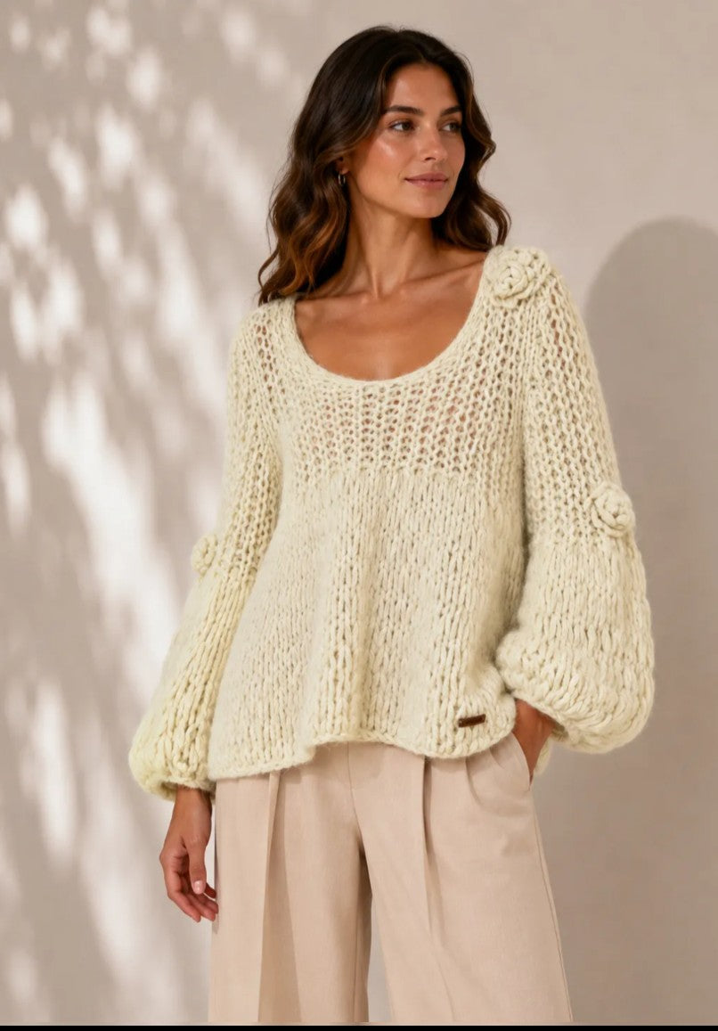 A woman wearing a chunky cream-colored open-knit sweater with balloon sleeves and beige wide-leg trousers, standing against a warm beige wall with soft leaf shadows.